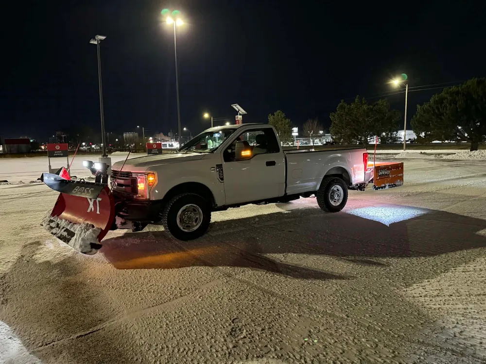 Plow truck with a red BOSS blade and rear pusher in a snowy lot at night